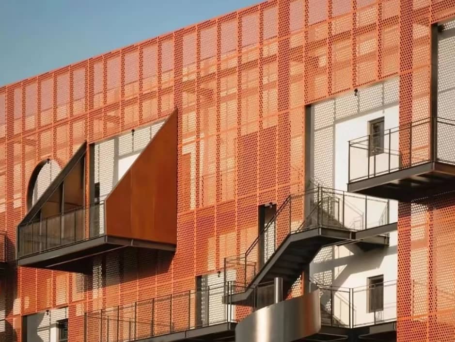 An orange perforated metal facade covers a modern building featuring black steel staircases and triangular balconies