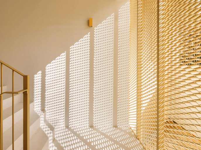 Warm sunlight filters through wooden blinds, casting striking diagonal shadow patterns across a cream wall. A golden-framed partition adds depth to this serene, minimalist interior bathed in soft amber tones