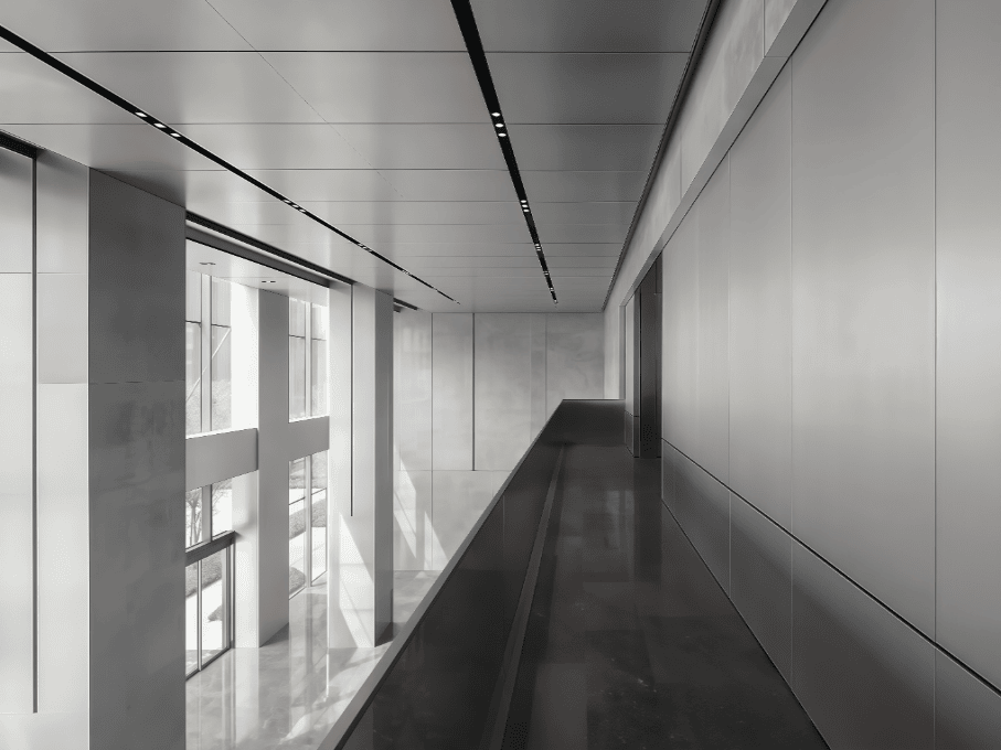 A minimalist modern corridor with sleek gray walls and polished dark floors. Linear black ceiling lights create dramatic lines leading to a bright window at the end, casting geometric shadows throughout the monochromatic space