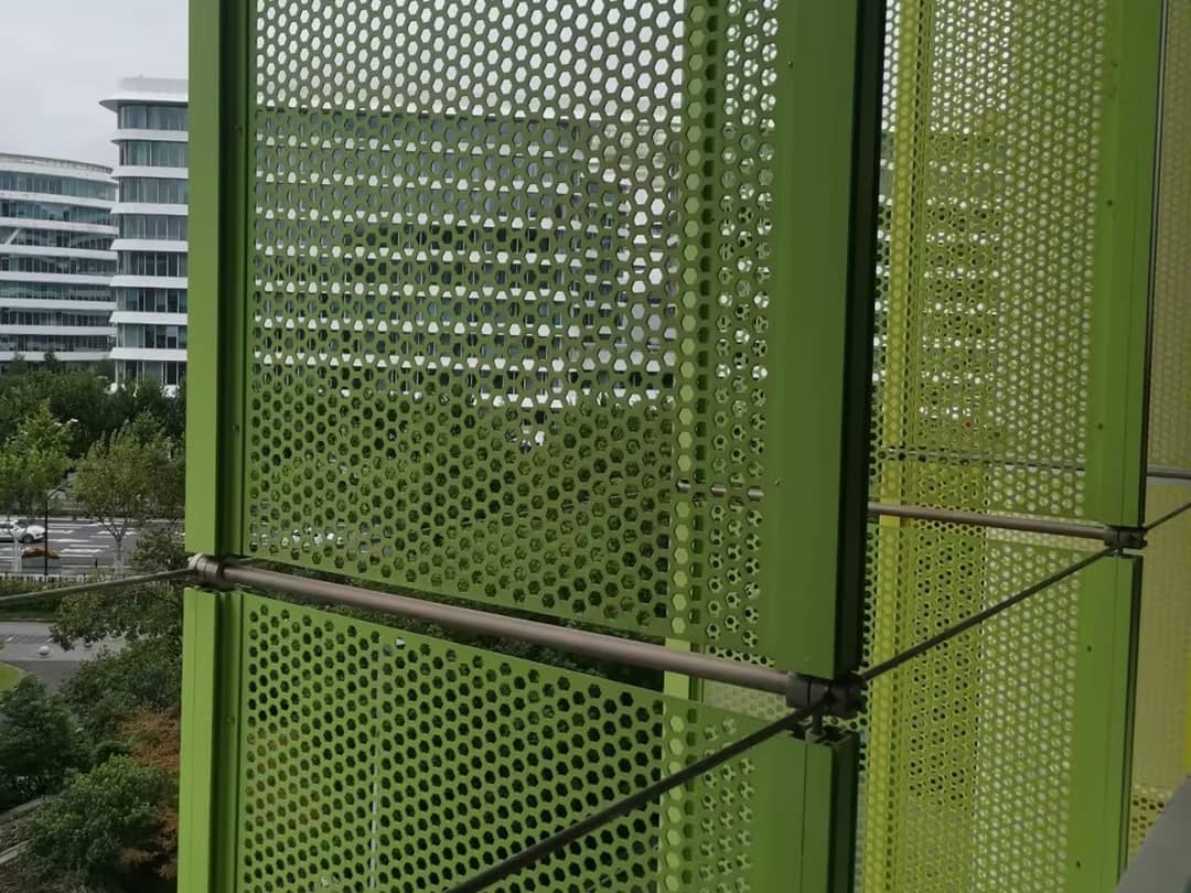 Green perforated metal panels are mounted on a building balcony overlooking a city street with white office blocks